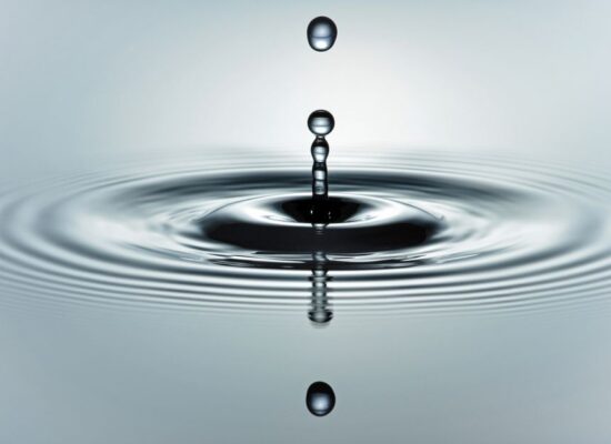 Close-up macro shot of a single water drop creating concentric ripples on a calm, serene water surface, symbolizing the ri...