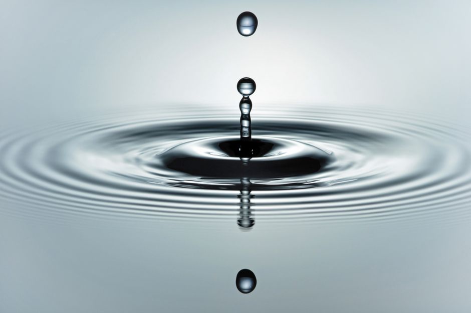 Close-up macro shot of a single water drop creating concentric ripples on a calm, serene water surface, symbolizing the ri...